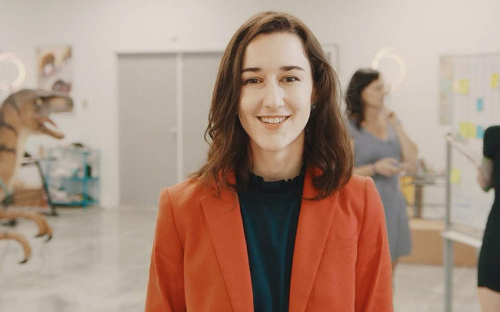Darby Cox in an orange blazer in the office, team working at a whiteboard behind her